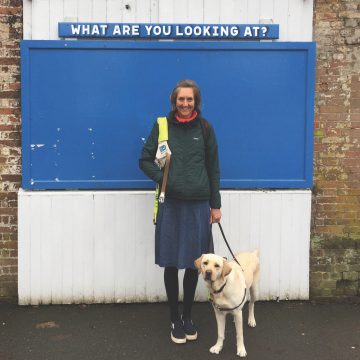 Angela Charles and Flynn. A photograph of a middle aged woman, the artist smiling at the viewer. She has light brown bobbed hair and wears a green jacket and a knee length denim skirt. She has a Guide Dog to her left, a yellow Labrador who is standing on all fours facing the viewer. The Guide Dog is on a lead and the women has the Guide Dog harness over her right shoulder. She is standing in front of a blue and white notice board with the words ‘What Are You Looking At’ along the top of the board above her head.