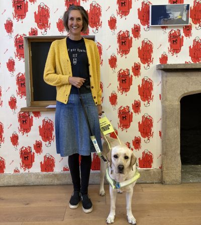 Review of Angela Charles Exhibition. A smiling middle aged woman with a short brown bob wears a bright yellow cardigan over a black t-shirt and denim skirt. She stands with her Guide Dog, a yellow Labrador in front of some white and red wallpaper, decorated with pictures of women and dog waste bins.